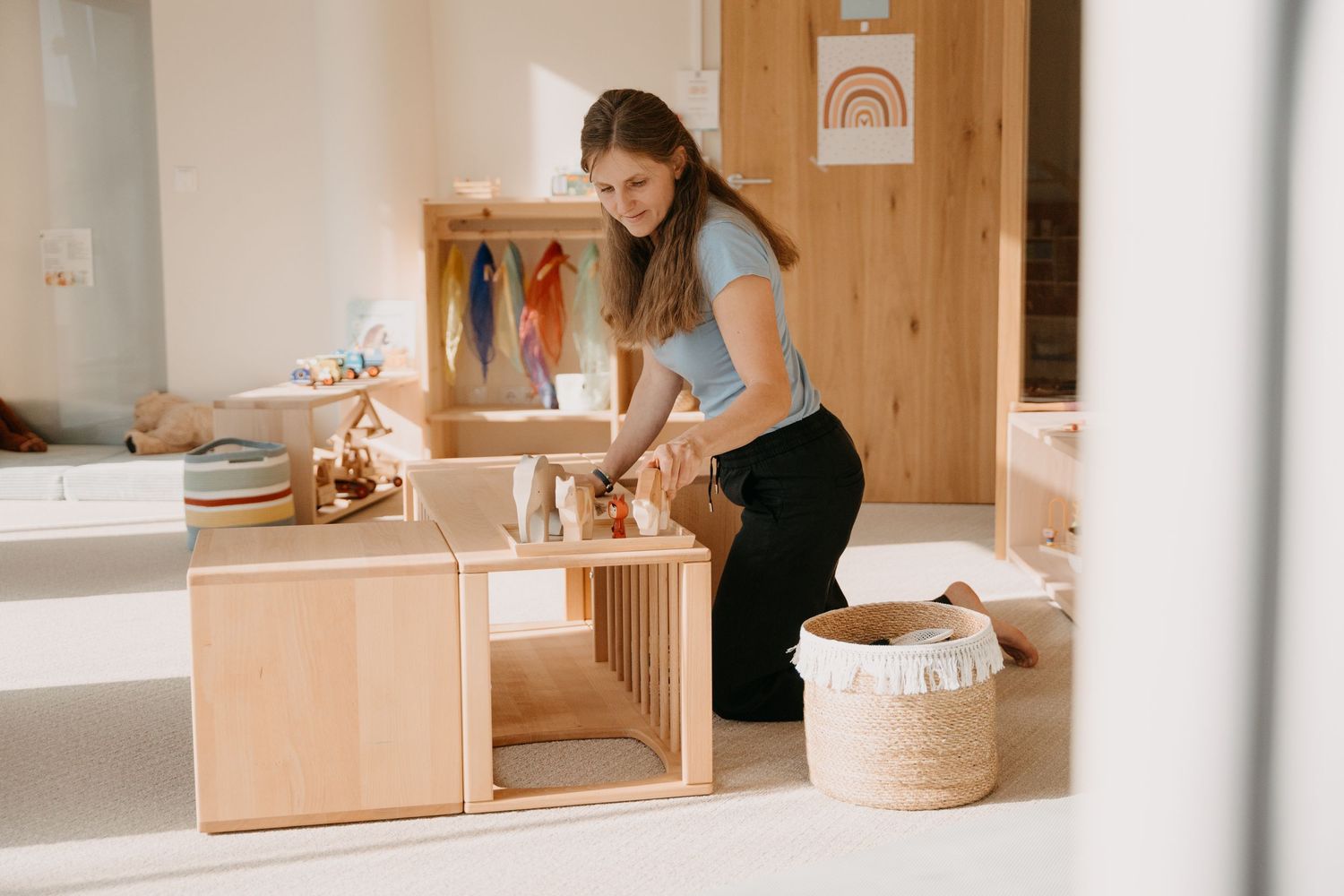 A person arranges wooden Montessori learning materials and toys on shelves in a bright, minimalist room.