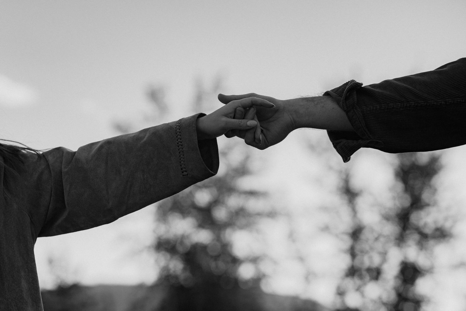 Two hands reach toward each other against a cloudy sky in a dramatic black and white composition.
