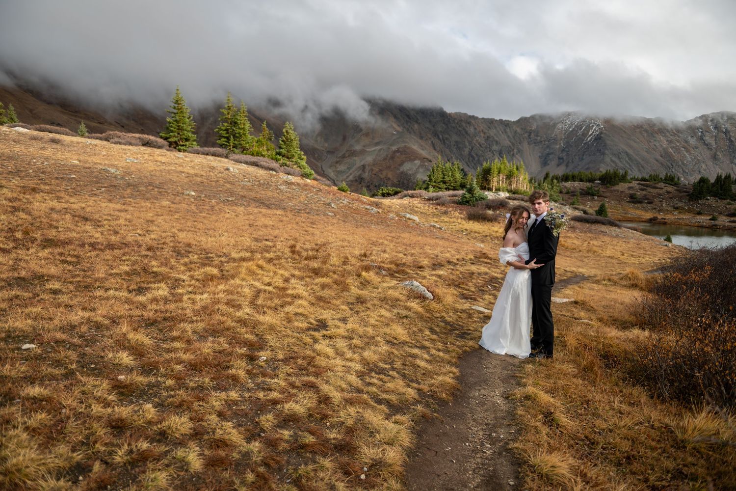 A bride and groom embrace on a mountain trail surrounded by golden meadows and dramatic cloudy peaks in an autumn elopement wedding photo.