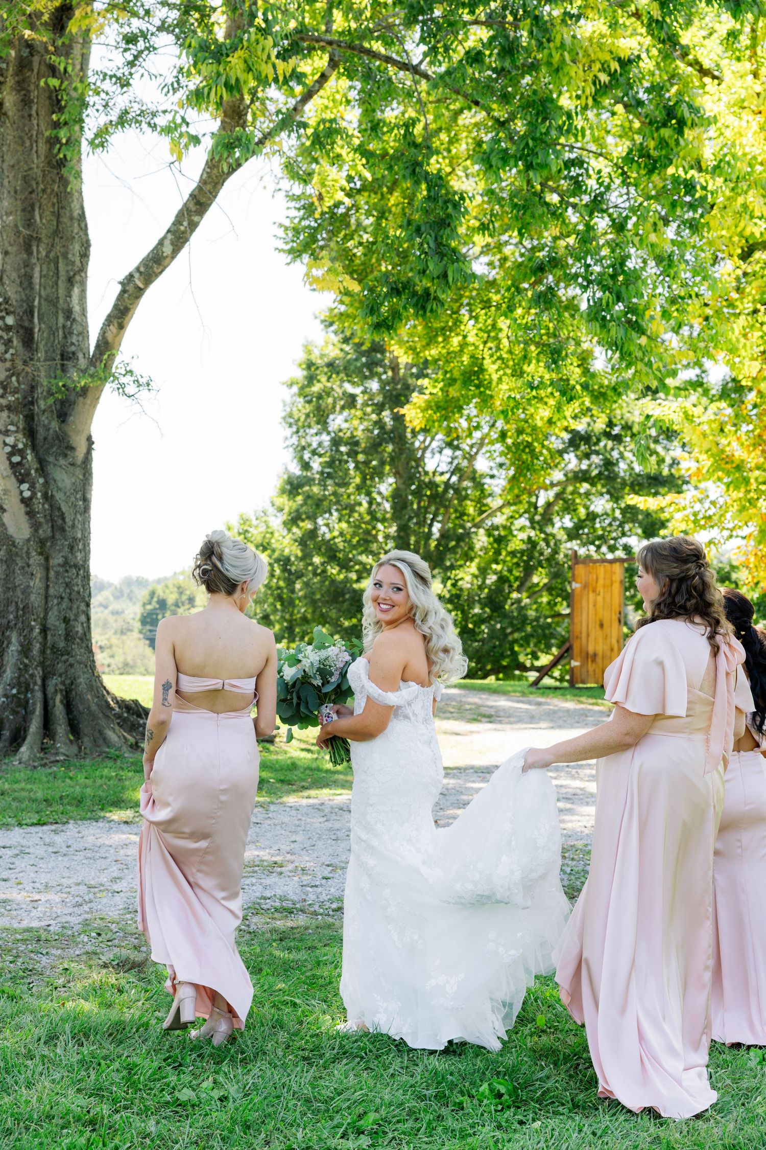 Bridesmaids in blush pink dresses help the bride with her wedding gown outdoors under green trees on a sunny day.