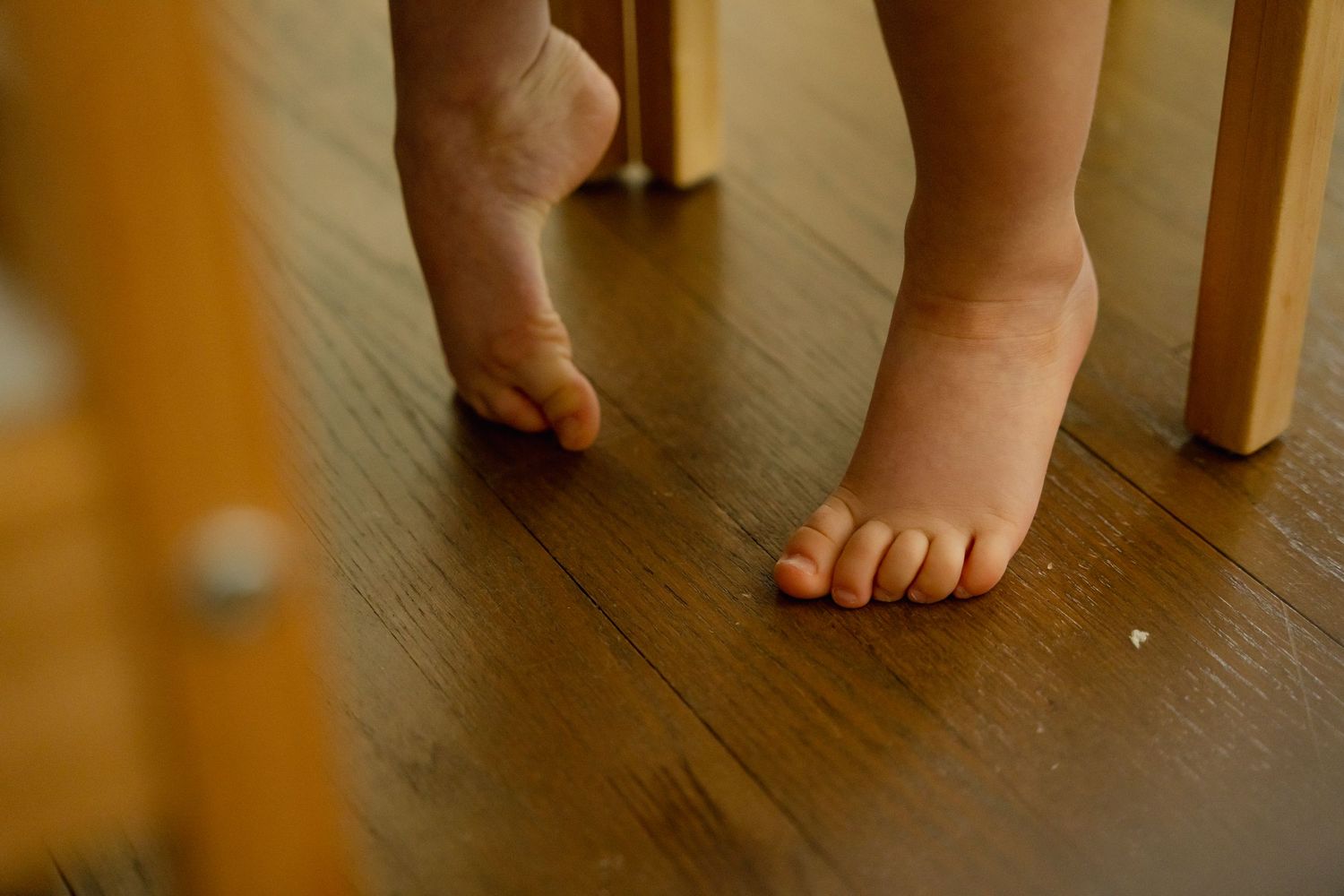 Bare feet standing on a hardwood floor next to wooden furniture legs in warm lighting at family photo session in Mount Prospect