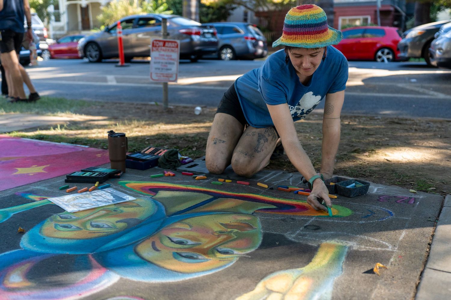 Artist creates vibrant chalk art design on pavement during outdoor festival in dappled sunlight.