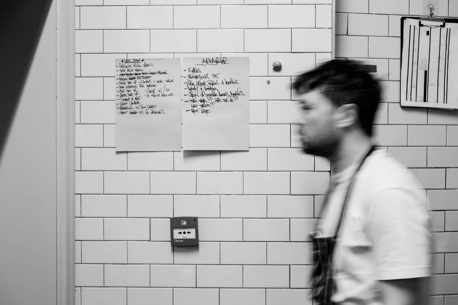 Black and white sequence of someone walking past a tiled wall with text and papers posted on it.