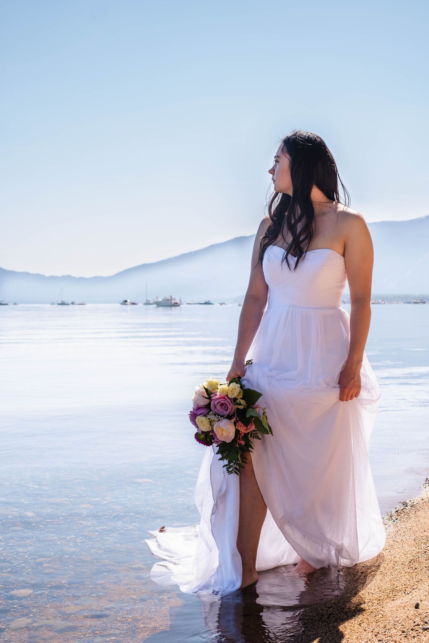 A bride in a flowing white dress stands at the water's edge holding a colorful bouquet against a mountain backdrop. Lak Tahoe CA