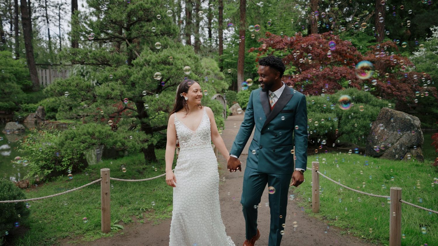 A couple in wedding attire walks hand in hand through a lush green garden path during their spring celebration.
