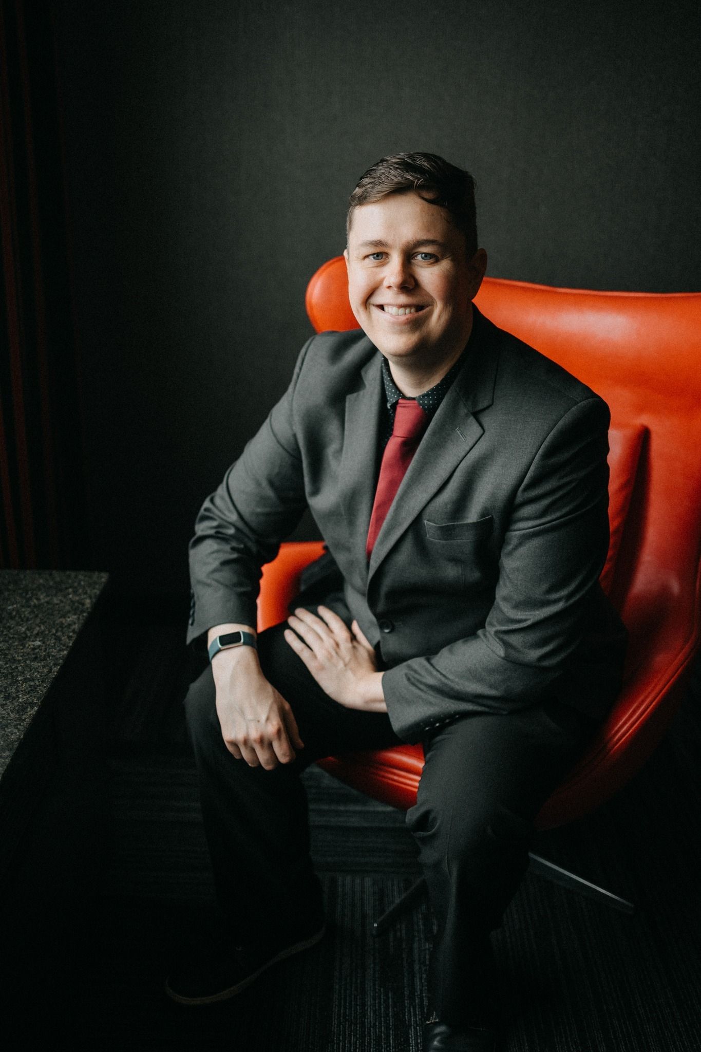 Professional studio portrait of someone in dark suit and red tie seated on red leather chair against black background.