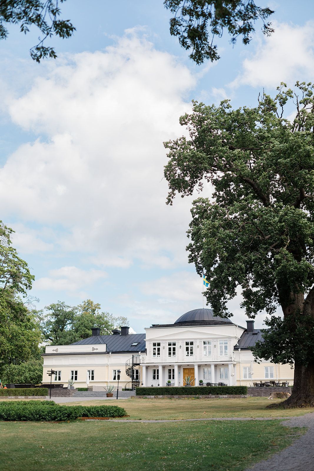 White single-story building complex with multiple wings surrounded by green lawn and mature trees on a sunny day.
