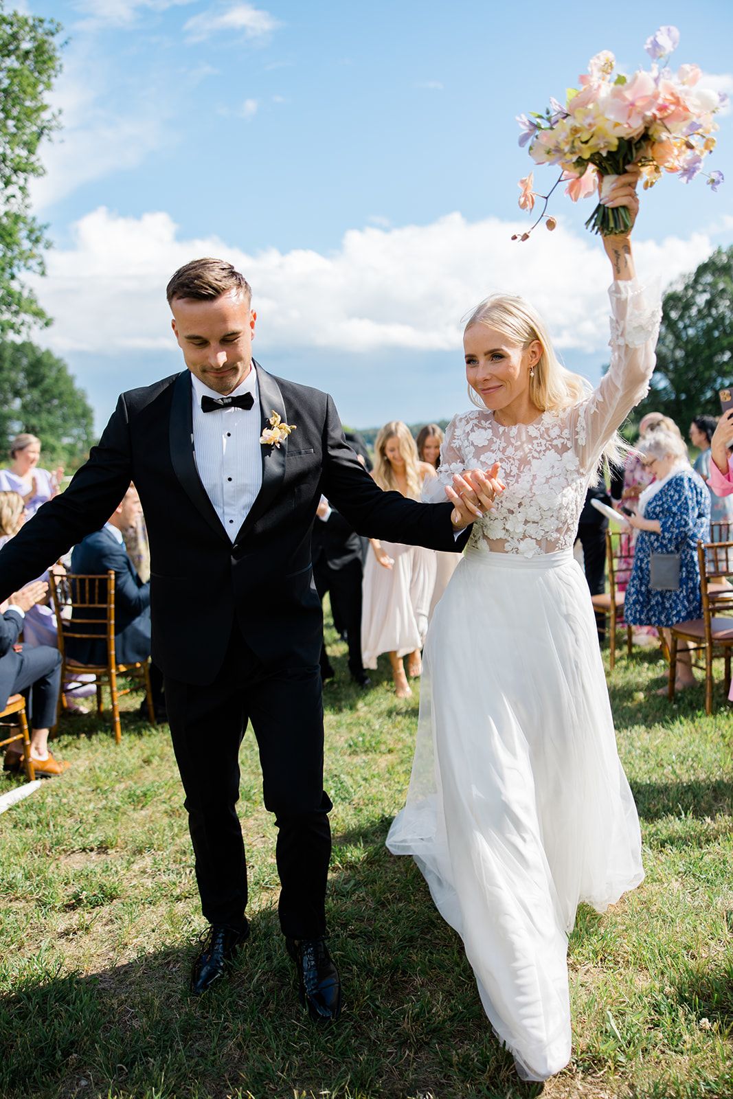 Newlyweds walk down a grassy hill during an outdoor wedding ceremony while guests throw flower petals in celebration.
