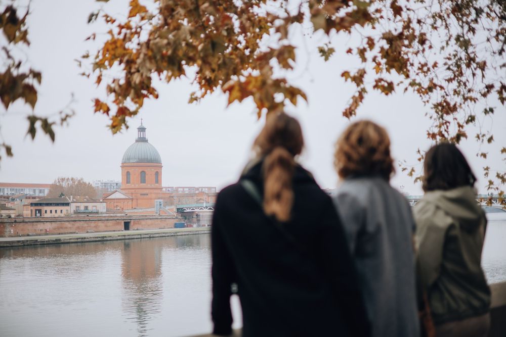Two people gaze at the domed Hospital de la Grave across the Garonne River in autumn Toulouse.