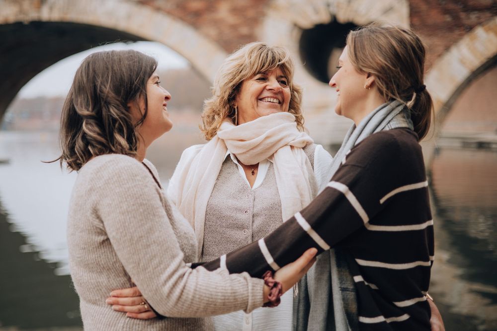Three women laugh and hold hands near a stone bridge arch along the Garonne River in Toulouse.