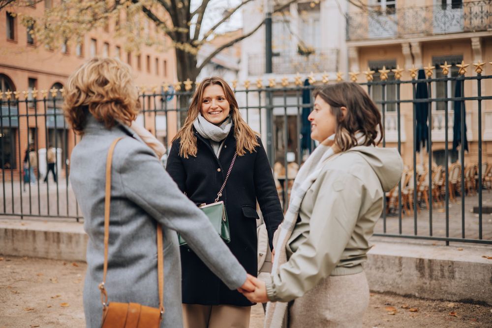 Three women in winter coats smiling and holding hands outdoors in a city park.
