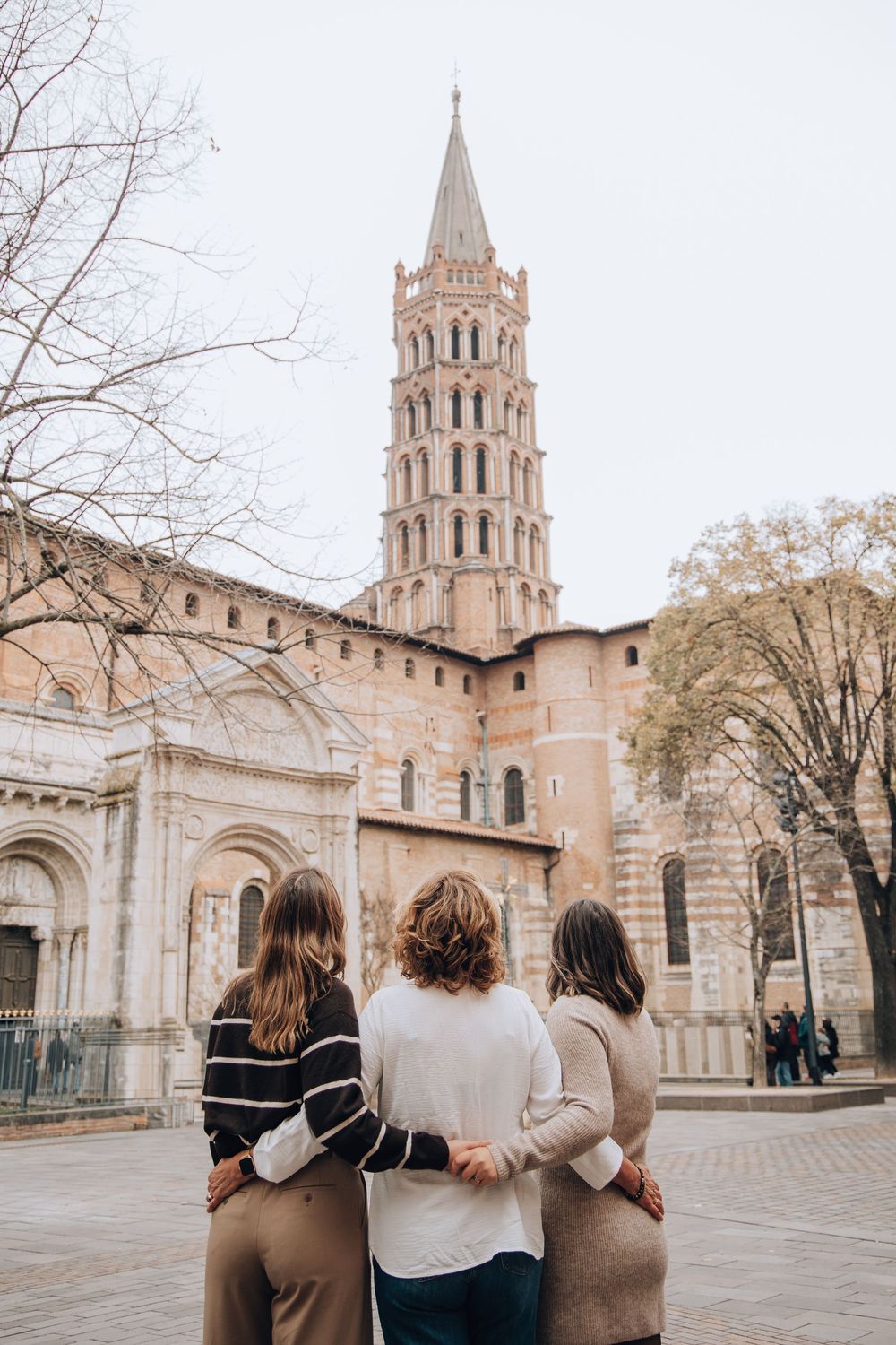 Three friends embrace while admiring the Basilica of Saint-Sernin tower in Toulouse, France.