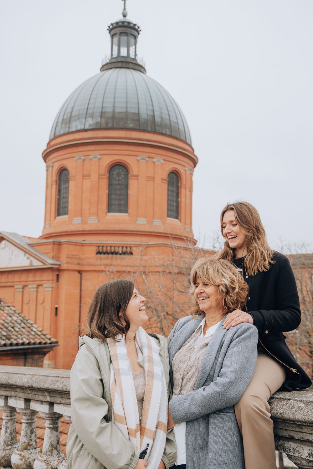 Three women laugh together in front of the iconic red brick dome of Hotel-Dieu Saint-Jacques in Toulouse.