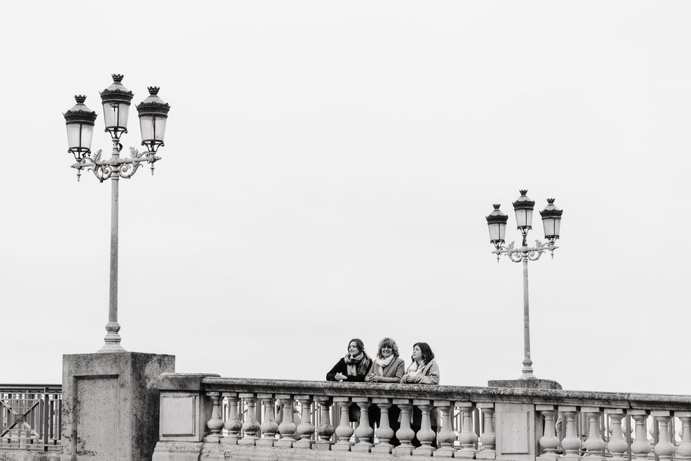 Three people stand between ornate lampposts on a classic stone bridge balustrade in Toulouse, France.