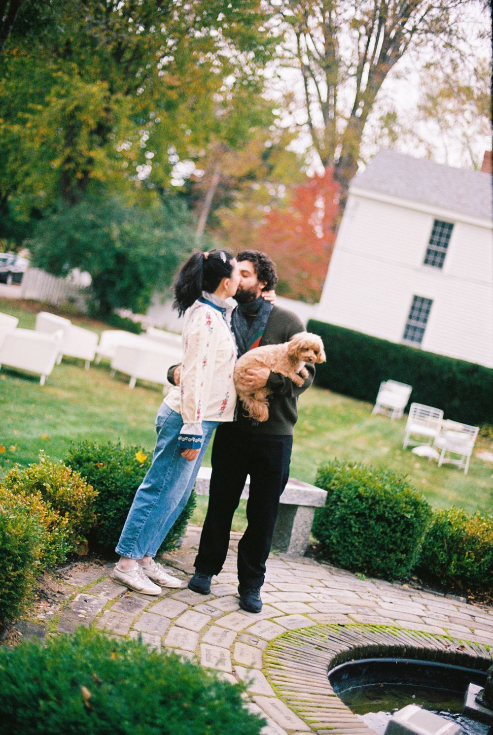 Couple kisses in the manicured garden by the fountain while he holds their dog in his arms and she wears fashionable maternity clothing.