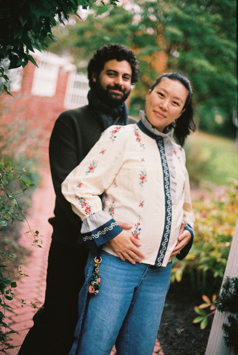 Husband stands behind his pregnant wife as they embrace and smile outside on a soft and overcast day in October in southern Maine.