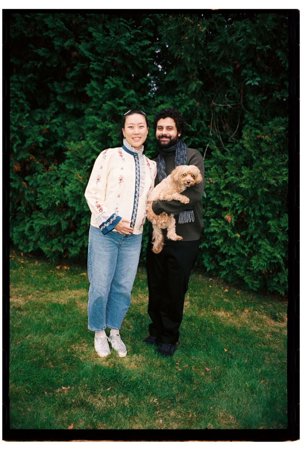 Couple embraces and pose together with their dog in the grass during a maternity family photoshoot on film in Kennebunkport, Maine.