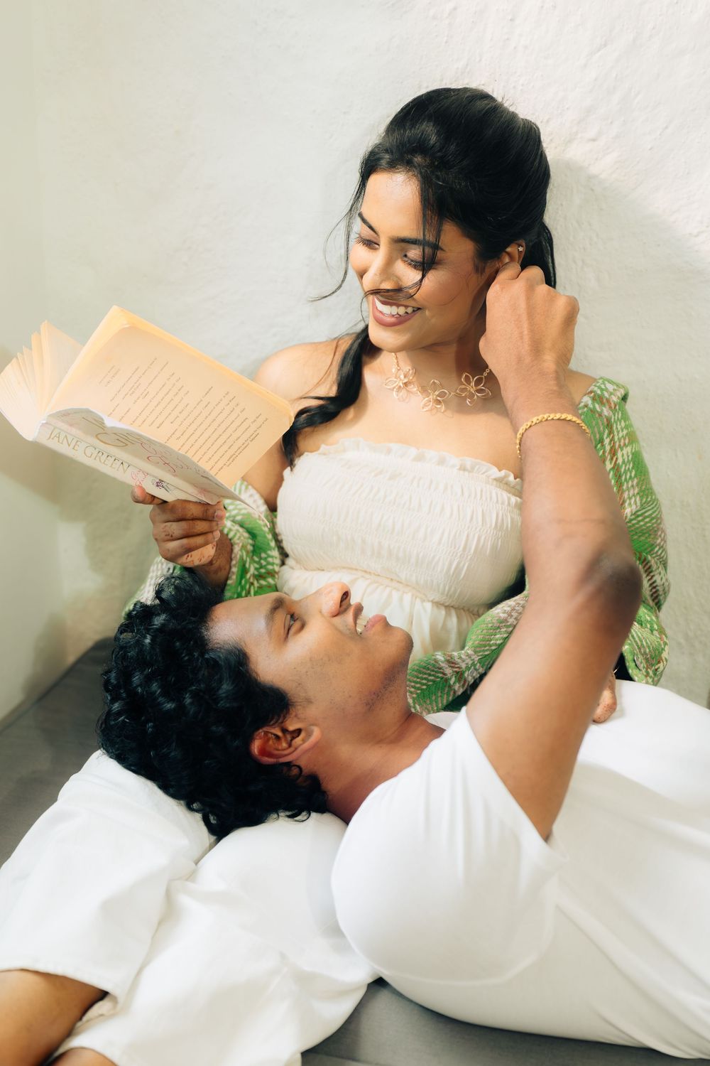 A couple shares an intimate moment as she reads a book while he rests his head on her lap on a couch.