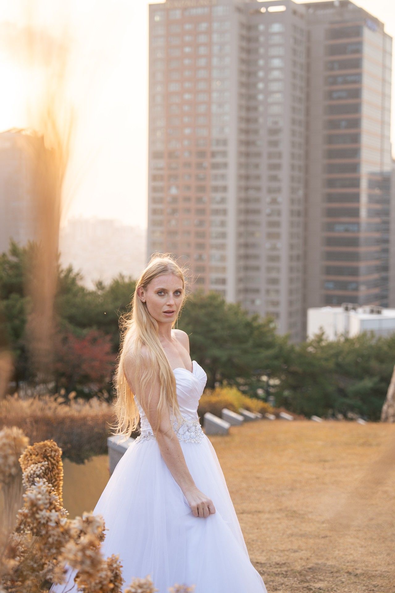 A girl in a white dress twirls gracefully in a field at sunset with city buildings in the background.
