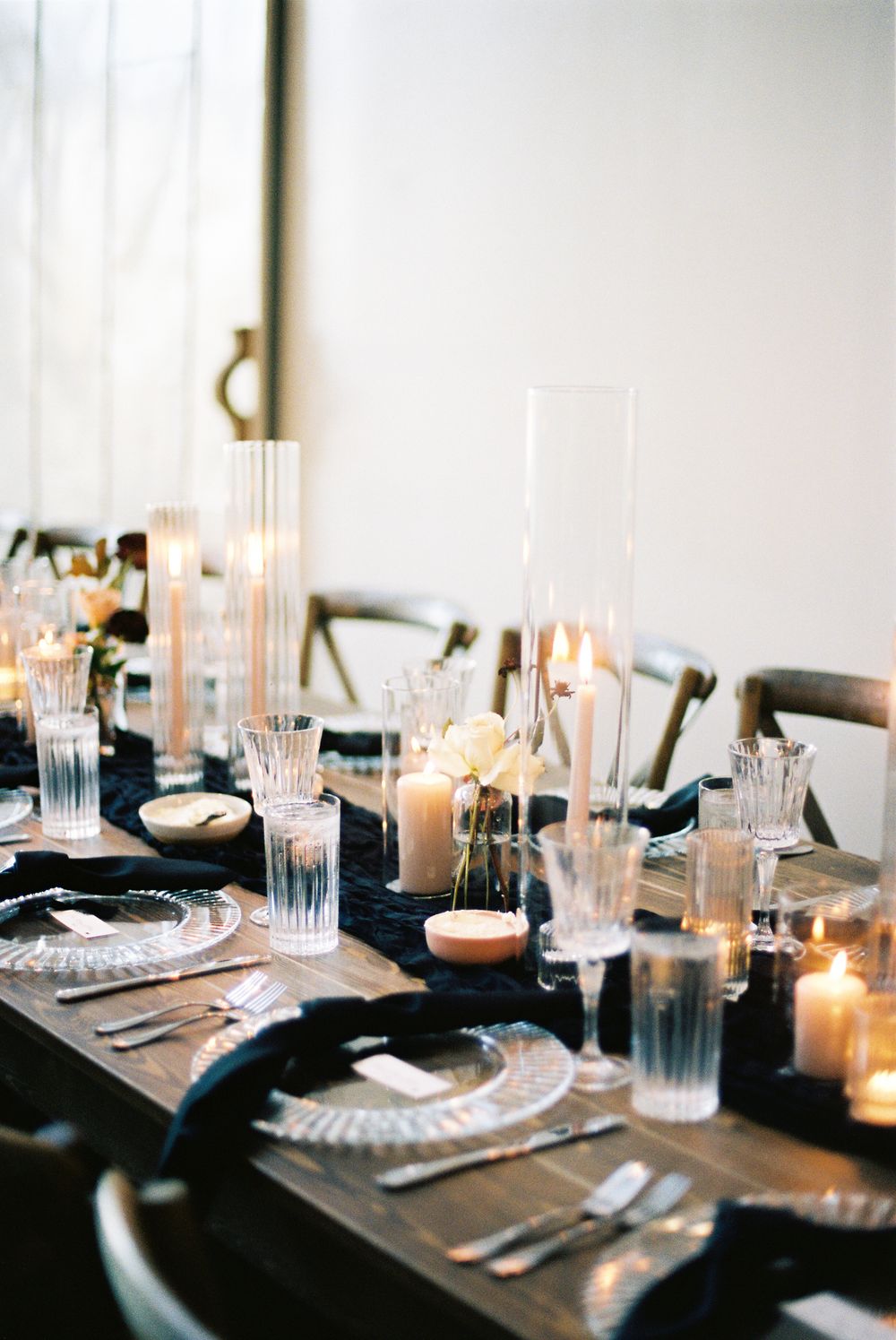 Elegant wedding reception table setting with glowing candles, crystal glassware and silver place settings on dark tablecloth.