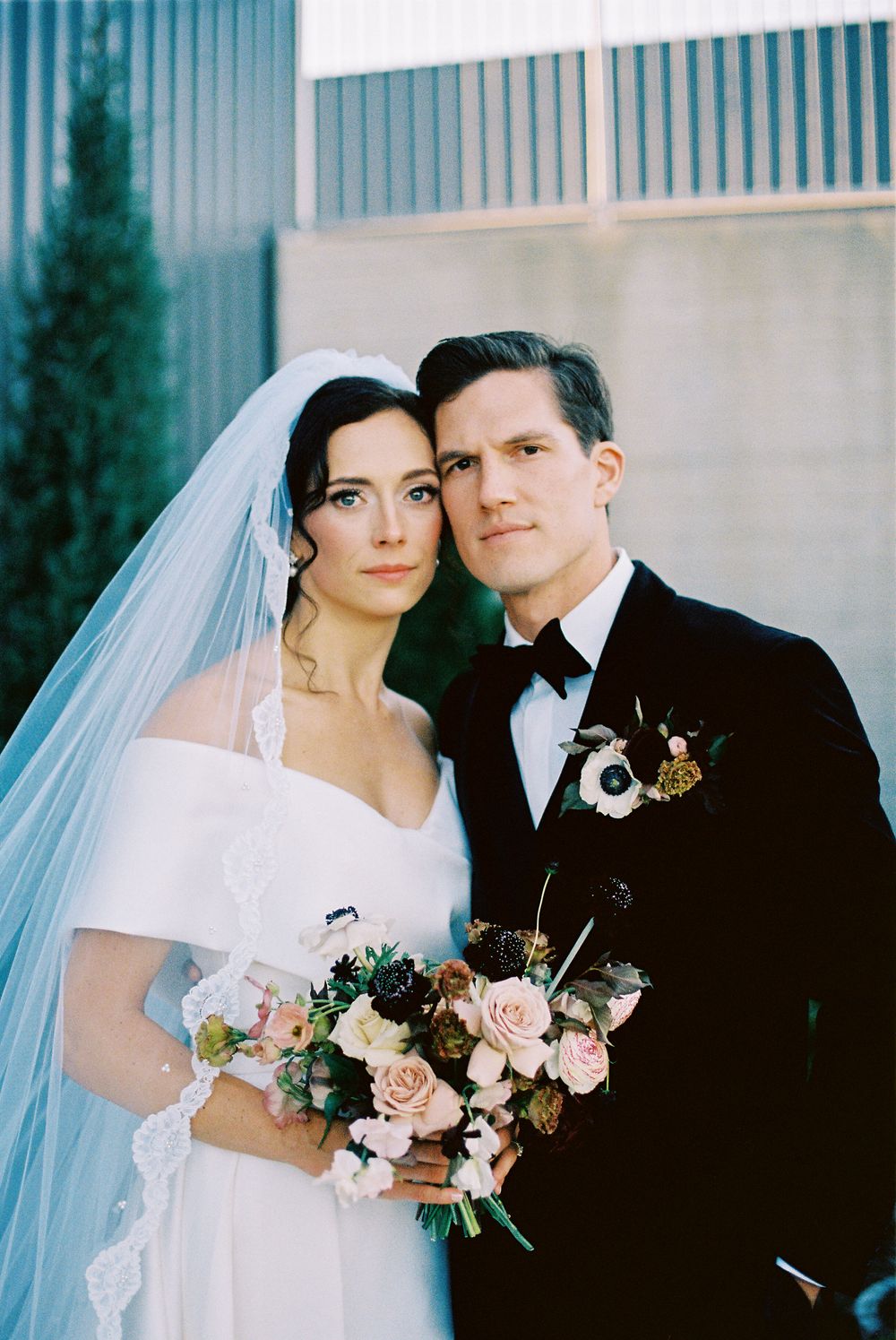 A bride and groom pose together in formal wedding attire with a bouquet of pink roses against a modern building.