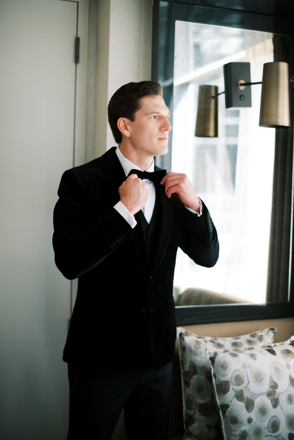 A person in a black suit gazes thoughtfully out of a window while standing in a modern interior space.