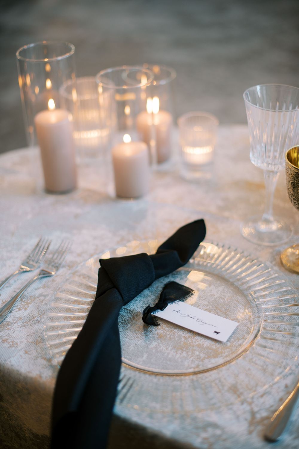 Elegant table setting with glowing candles, crystal glassware, and a black napkin on a white tablecloth.