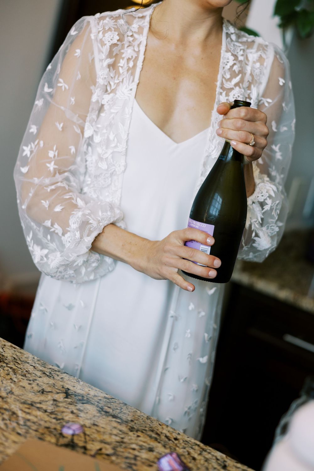Woman in white lace robe holding champagne bottle against marble counter.
