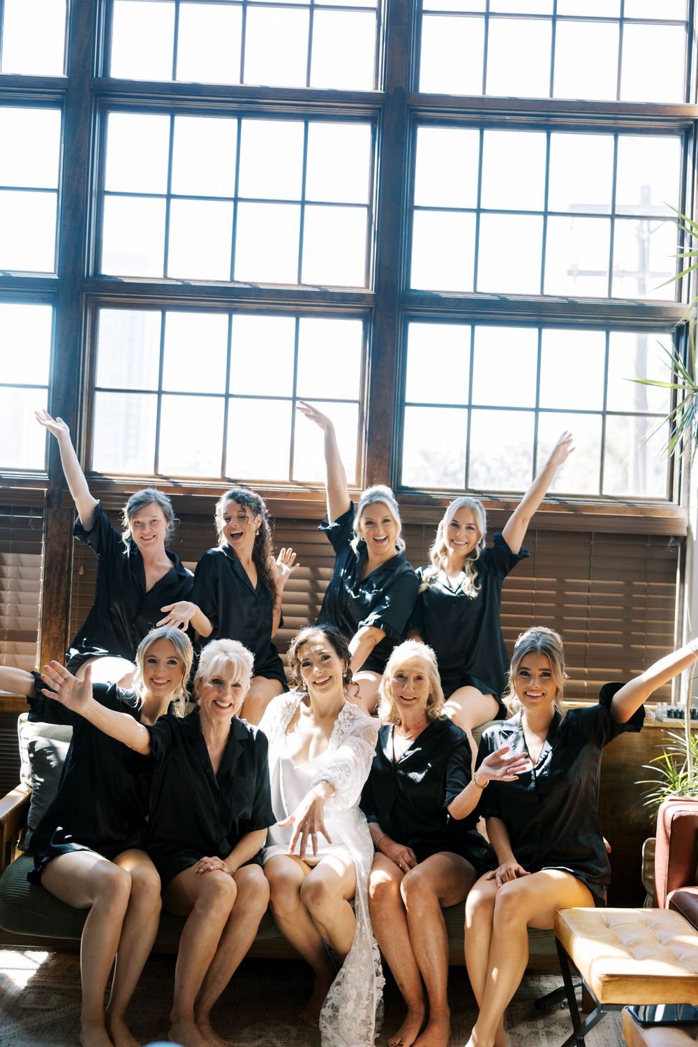 A group of bridesmaids in black leotards sit together on a bench, raising their arms in celebration.