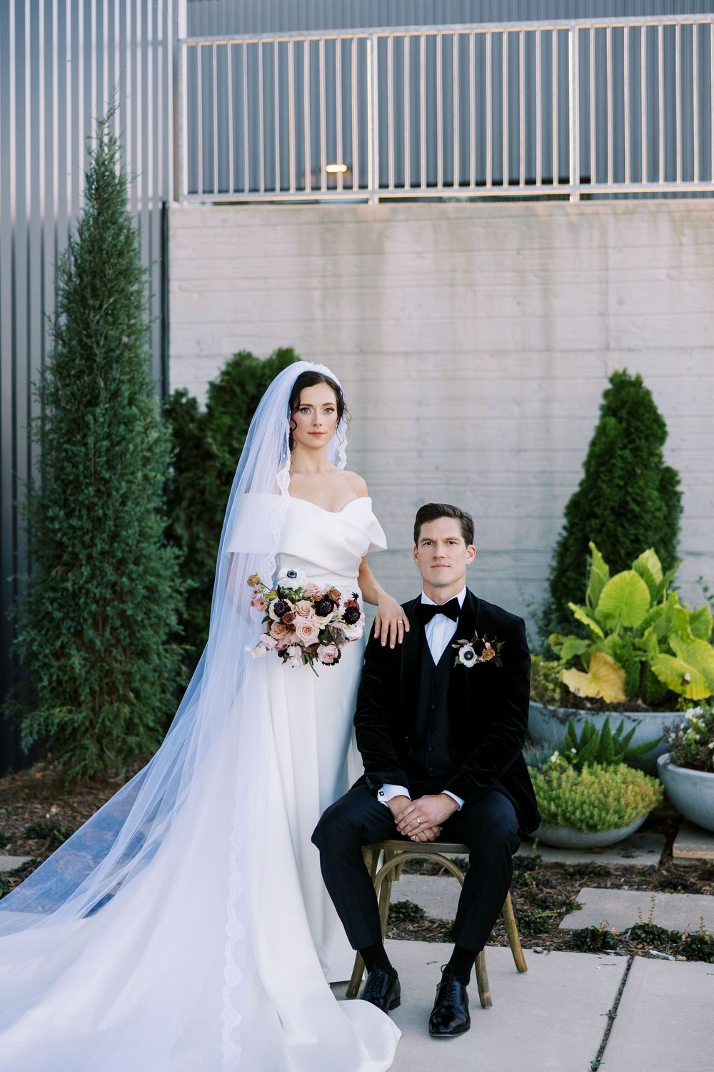 A wedding couple poses against a modern architectural backdrop with greenery and a long flowing veil.