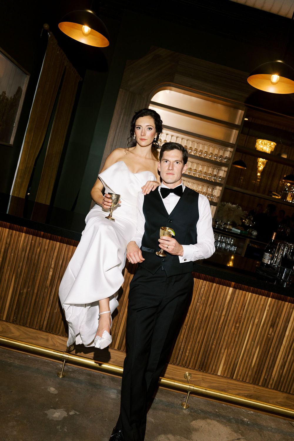 A couple in wedding attire holding drinks while posing in an elegant bar setting with warm lighting.