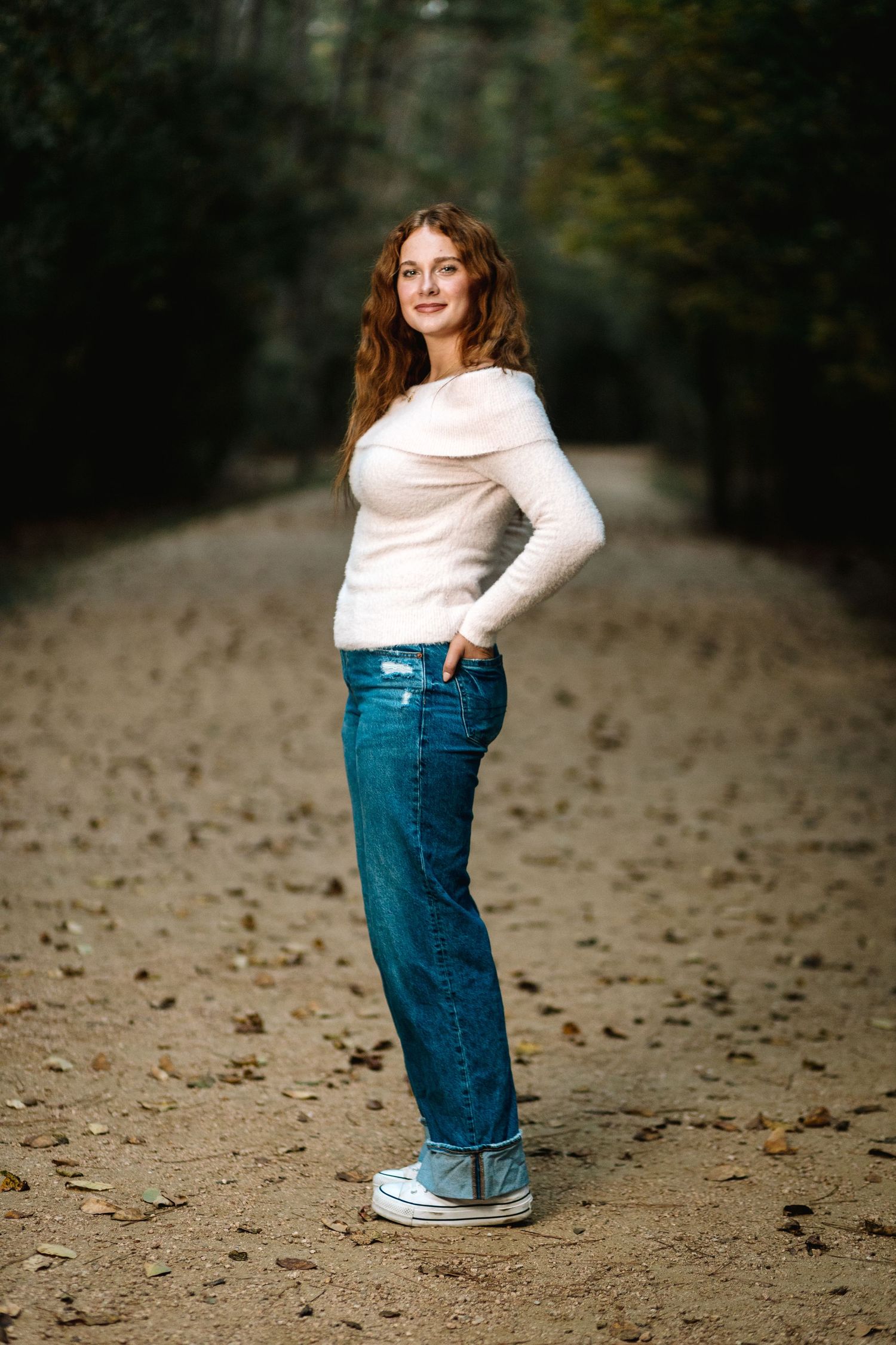 A casual portrait on a dirt path in the woods featuring a white sweater, blue jeans and sneakers.