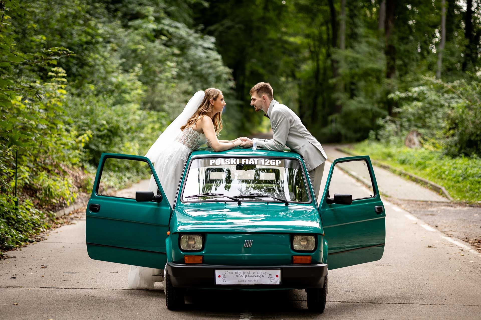 Wedding couple poses with a classic teal Fiat car in a forest setting.