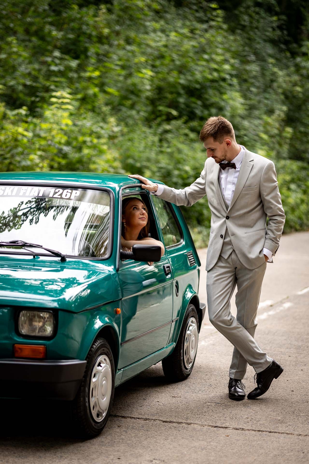 Wedding couple interacts playfully around a classic teal car on a forest road.