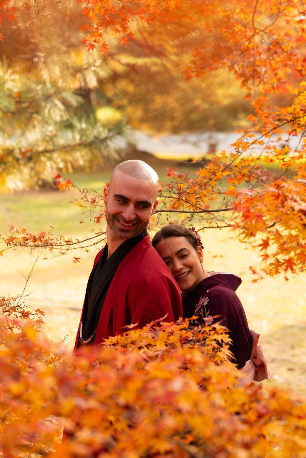 Smiling couple in traditional red and purple kimono embracing among vibrant orange autumn maple leaves.