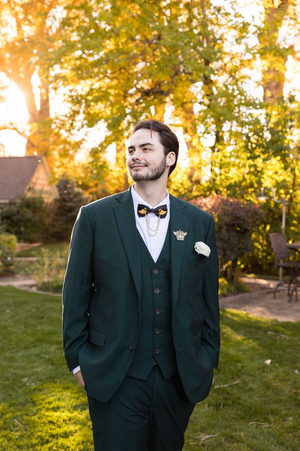 A person in a dark green three-piece suit and bow tie stands in a garden during golden hour sunset.