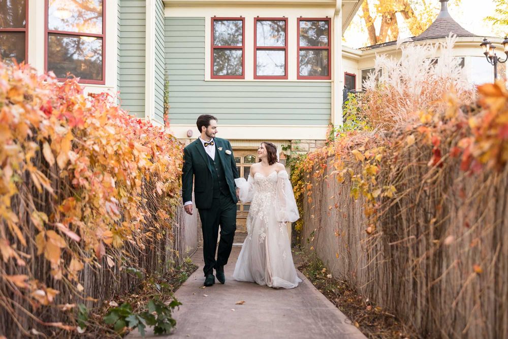 Couple in formal wedding attire walking through autumn foliage in a garden setting.