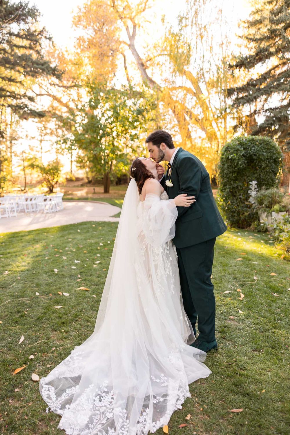 A couple in wedding attire shares a romantic moment outdoors during golden hour with fall foliage in the background.