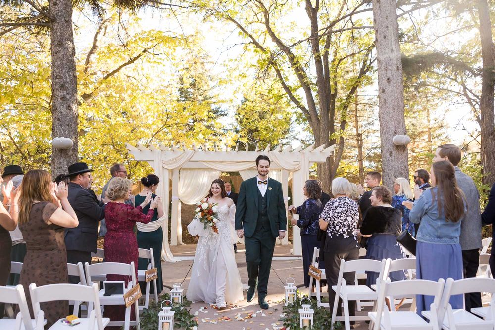 Wedding guests mingle outdoors at a fall reception with white chairs and golden autumn foliage in the background.