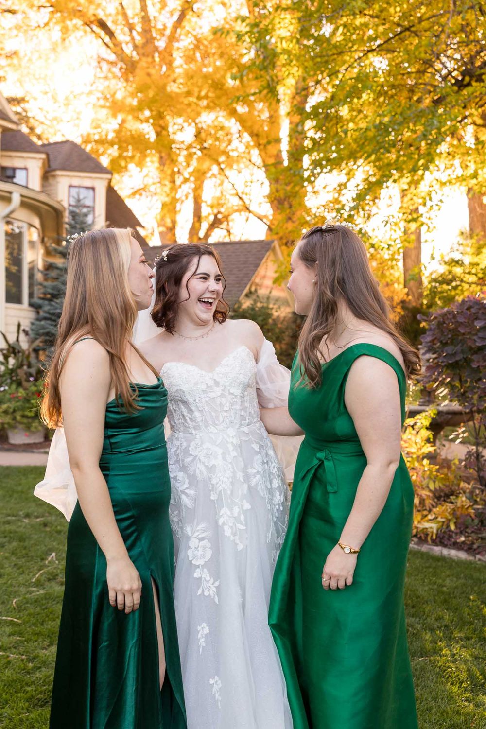 Wedding party in emerald green bridesmaid dresses and white lace gown share joyful moment at sunset under autumn trees.