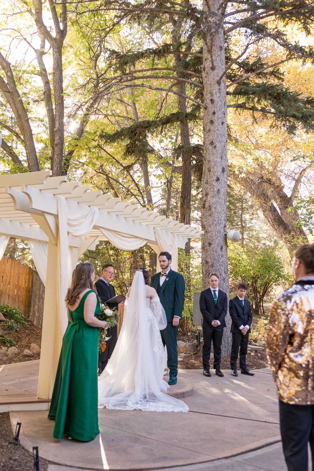 Wedding ceremony taking place under white pergola in wooded outdoor setting with guests in formal attire.