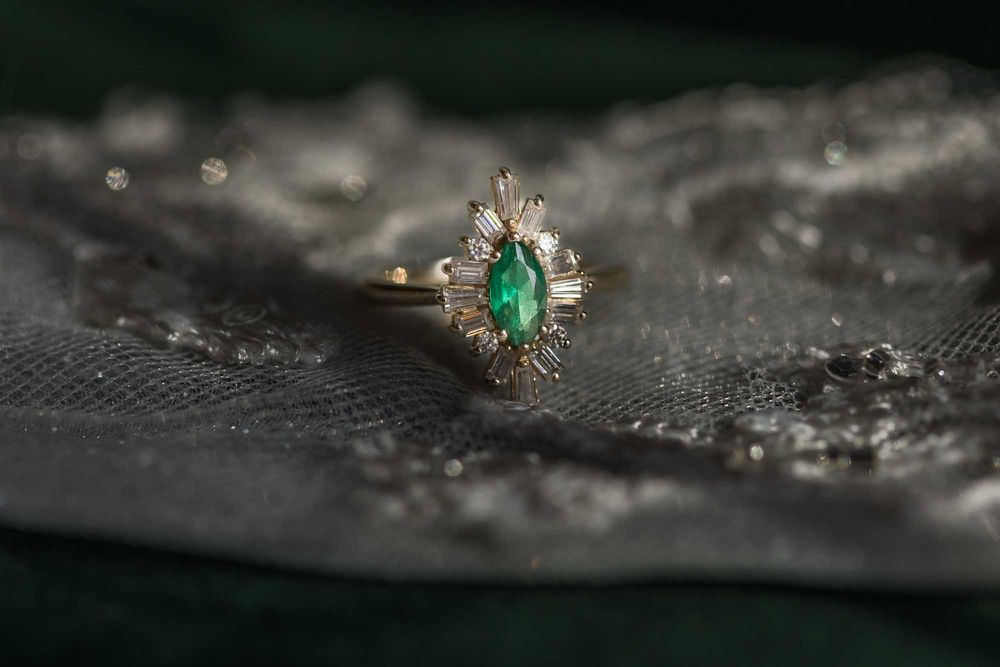Macro shot of emerald and diamond engagement ring with intricate details against dark textured background.