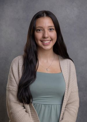 Professional portrait of person in sage green top and beige cardigan against gray backdrop.