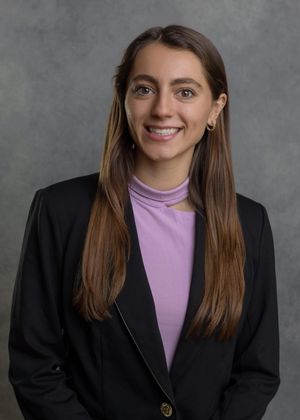 Professional portrait of person in black blazer and purple turtleneck against gray backdrop.