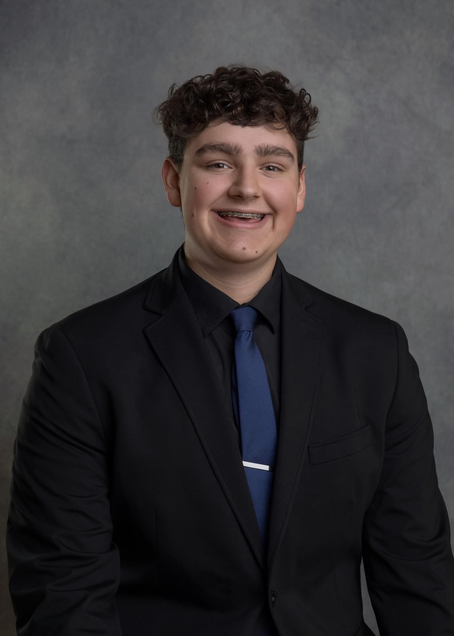 Professional headshot of person in dark suit and blue tie against gray background.