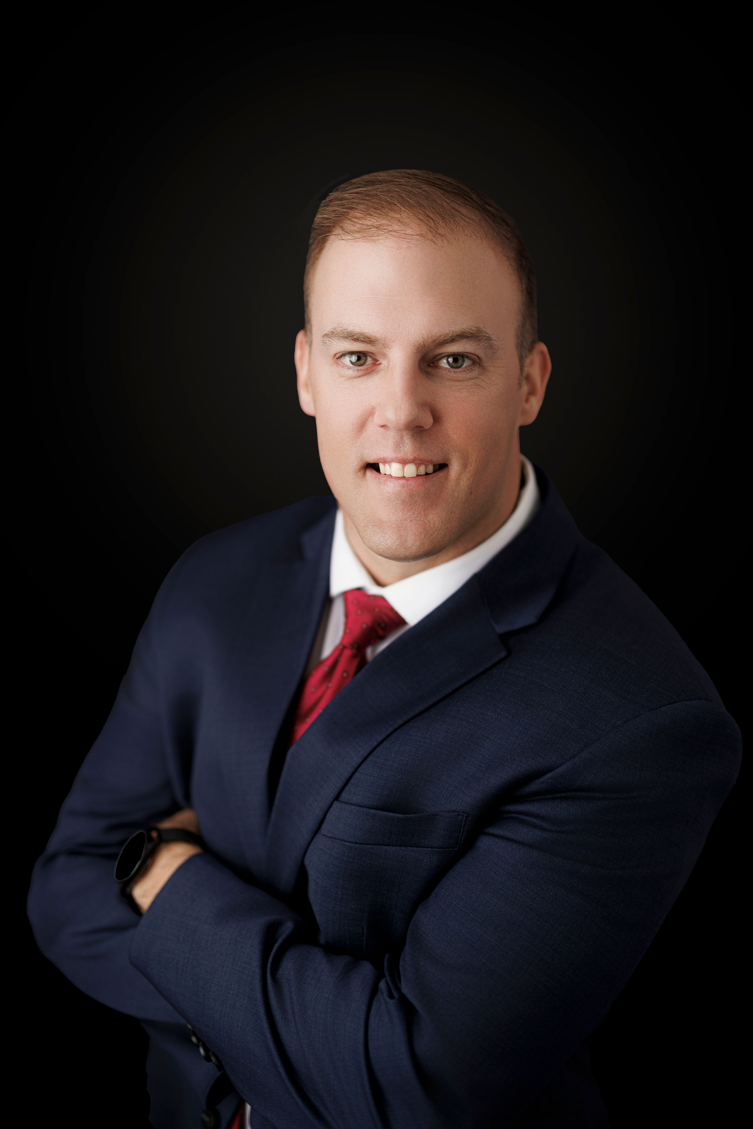Professional headshot portrait of someone in a navy suit and red tie against a dark background.