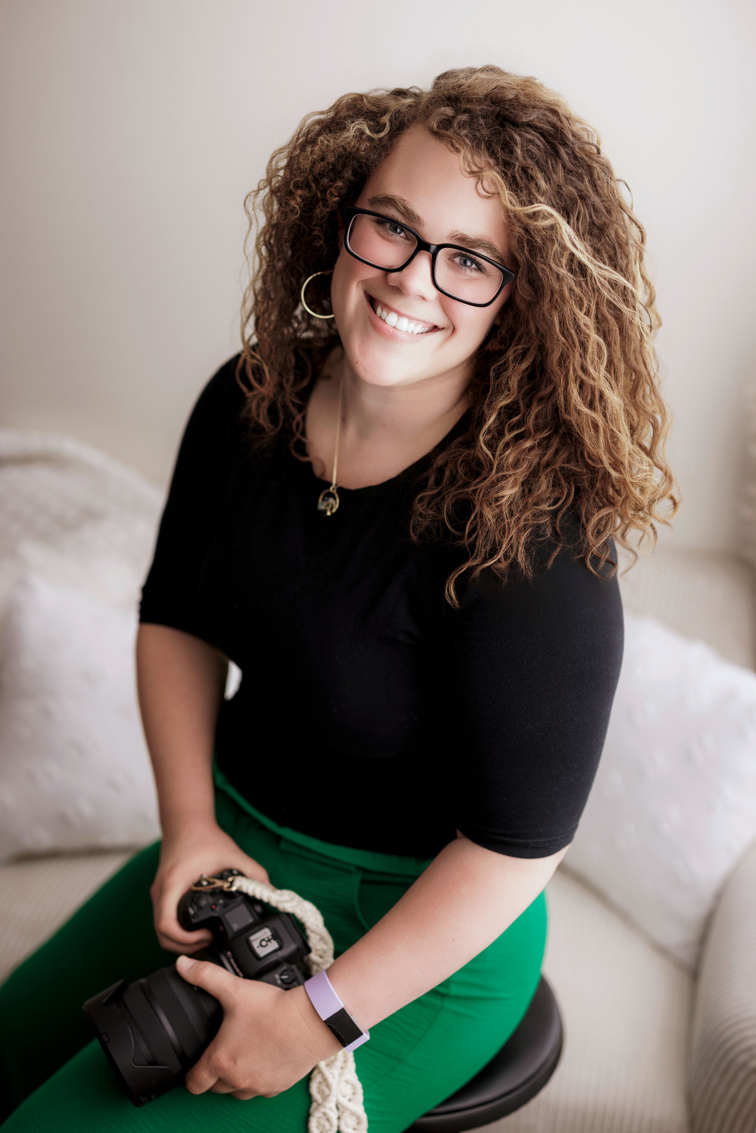 Professional photographer in black top and green skirt sits casually with camera gear on light colored couch.
