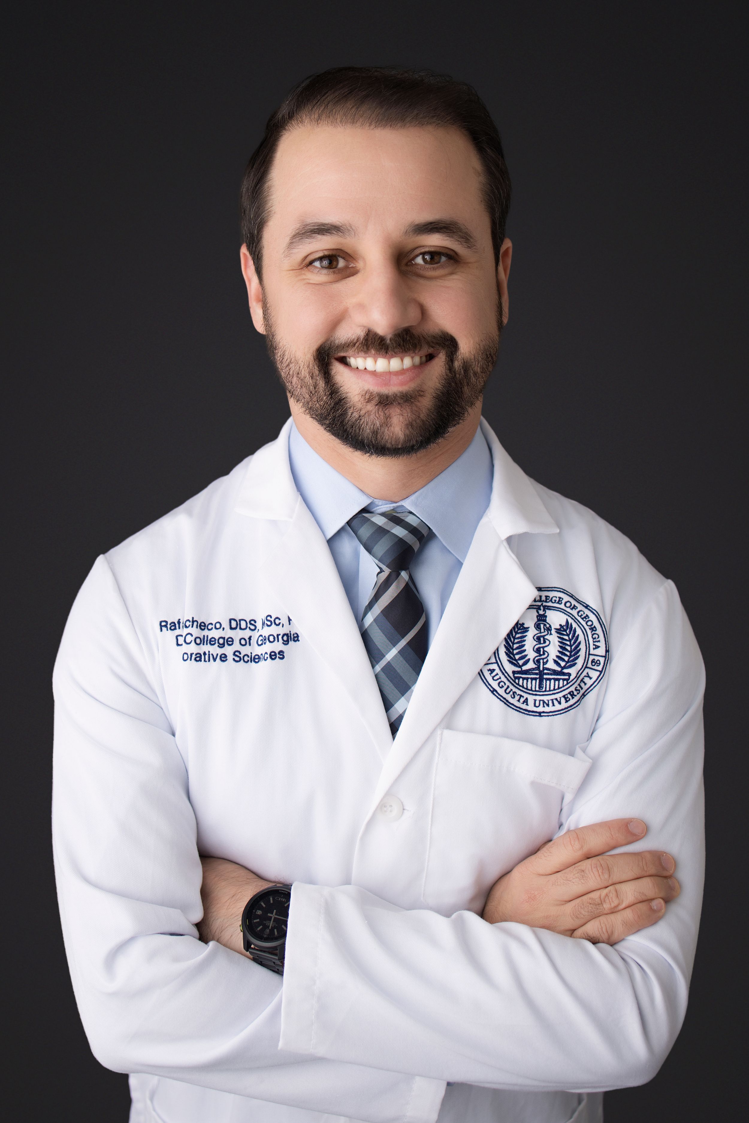 Professional medical staff member in white coat and striped tie poses with crossed arms against dark background.