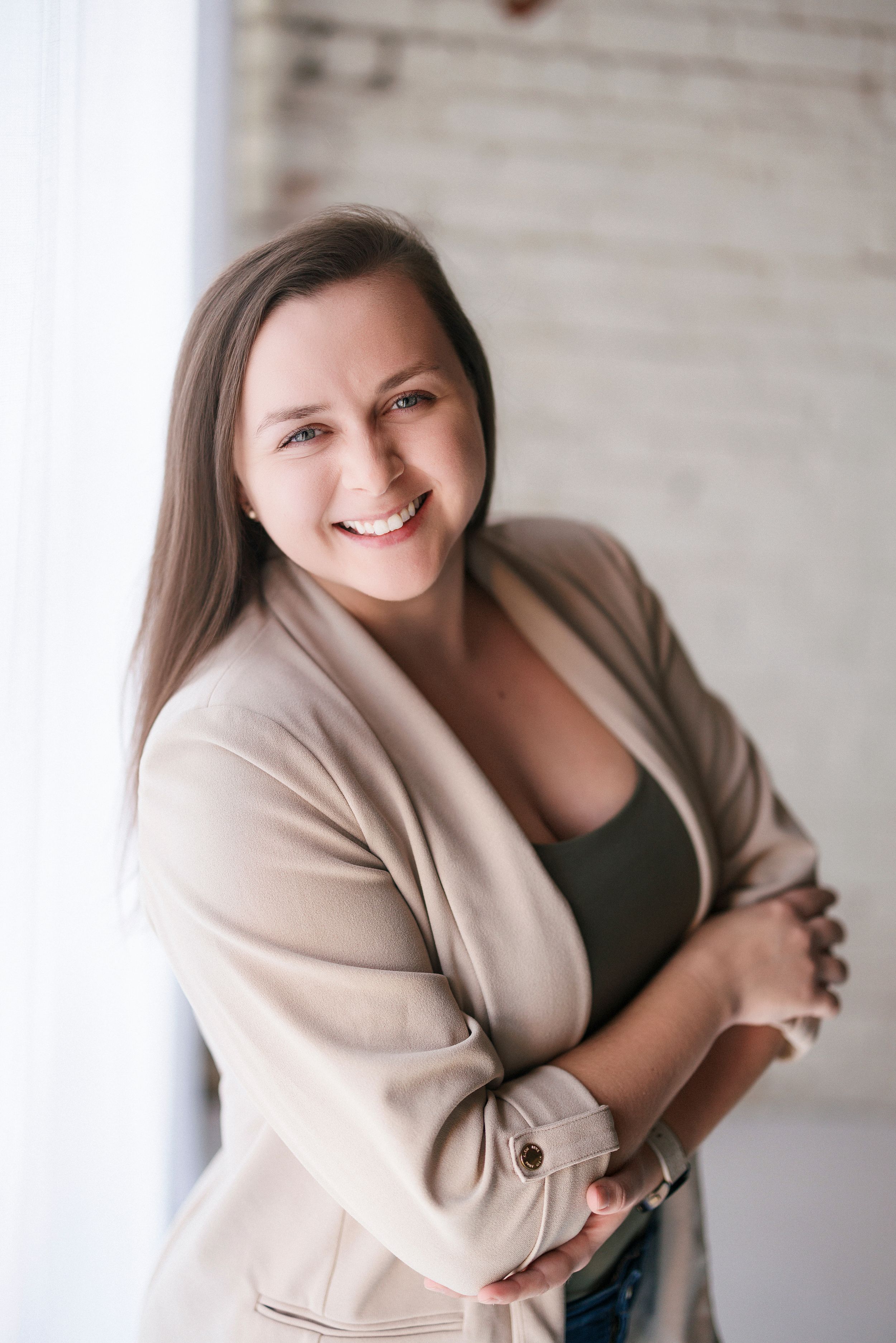 Professional portrait photo showing someone in a beige blazer and dark top against a light colored wall.