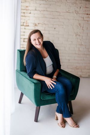Professional in navy blazer and jeans sits casually in teal armchair against white brick wall in modern studio setting.